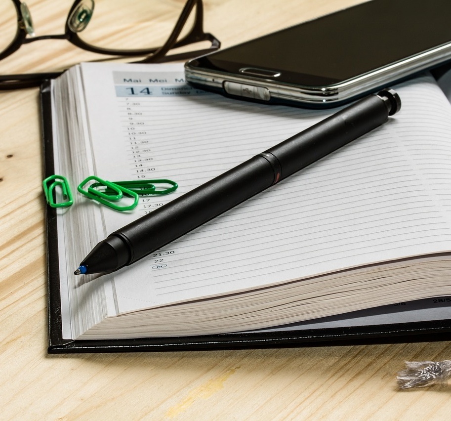 Ein Kalender, ein Stift, ein Handy und eine Brille liegen auf einem Tisch.
