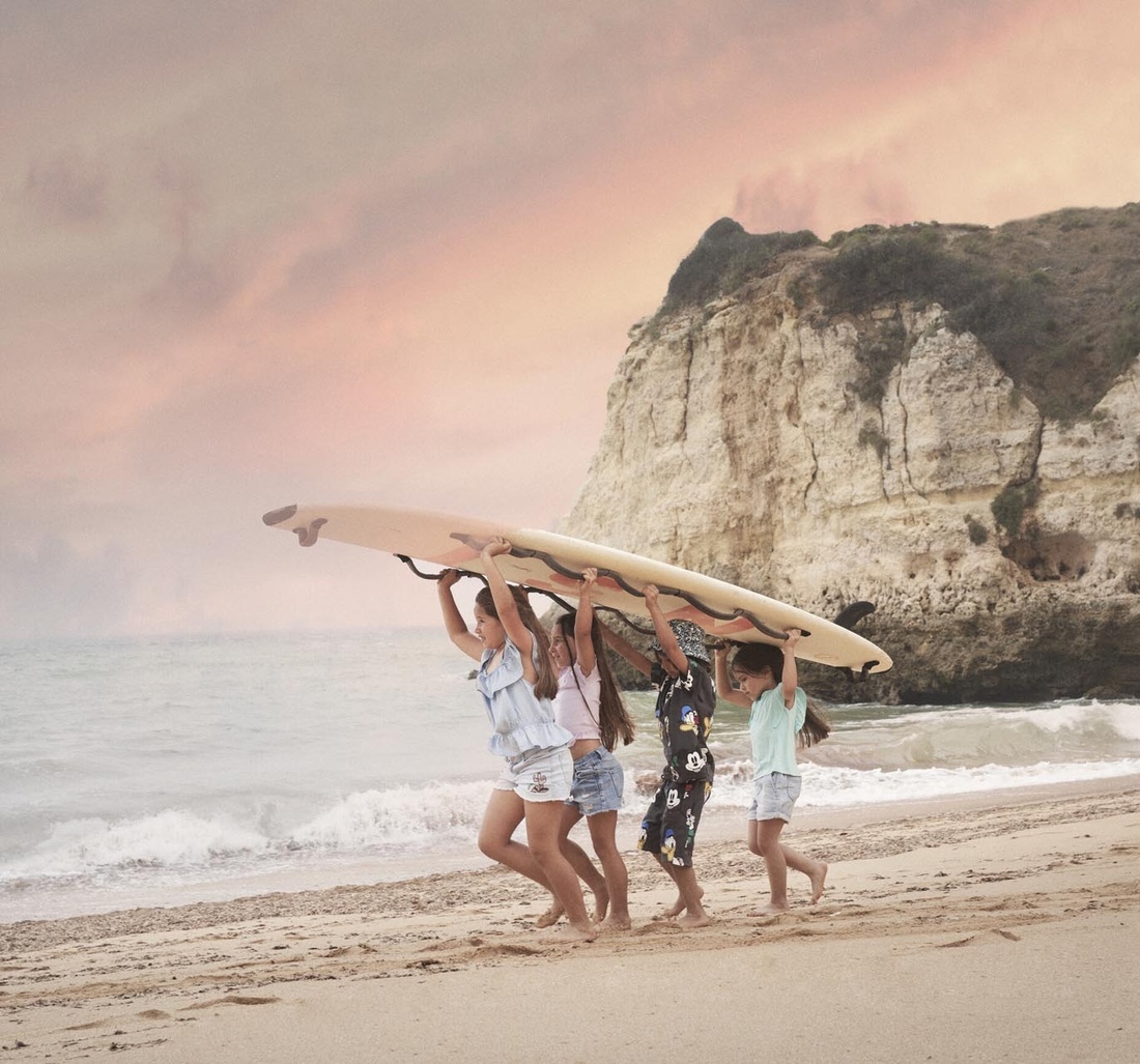 Vier Kinder tragen ein Surfboard am Strand.