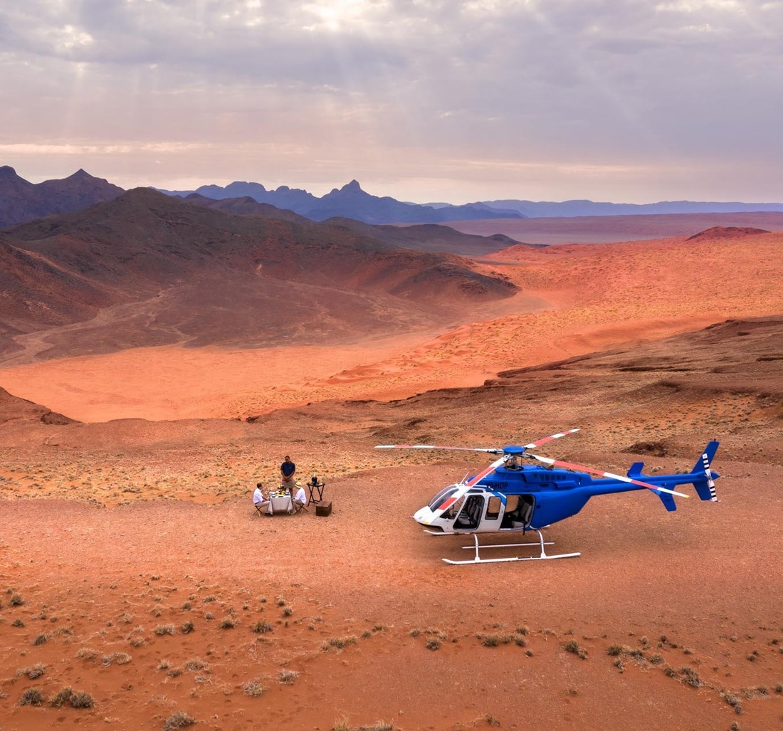 Ein blauer Helikopter steht, in der Wüste, neben drei Personen.