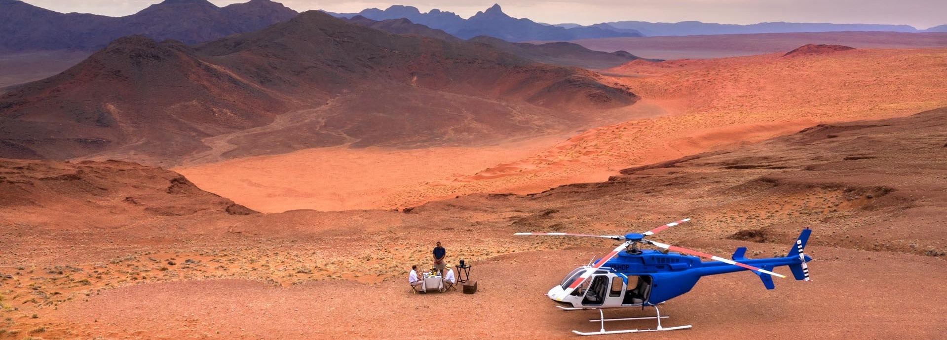 Ein blauer Helikopter steht, in der Wüste, neben drei Personen.